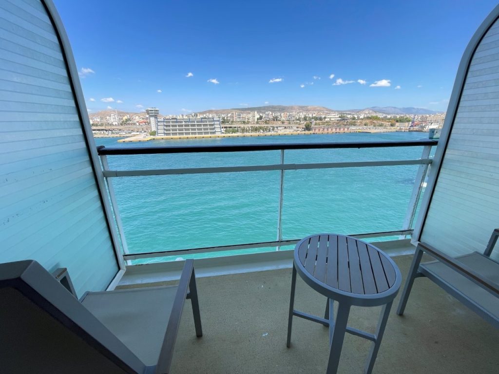 Two chairs and a small round table sit on the private veranda overlooking vivid turquoise water and the shoreline beyond. The balcony added valuable outdoor space and a private place to enjoy port views during the Celebrity Infinity Greece cruise.