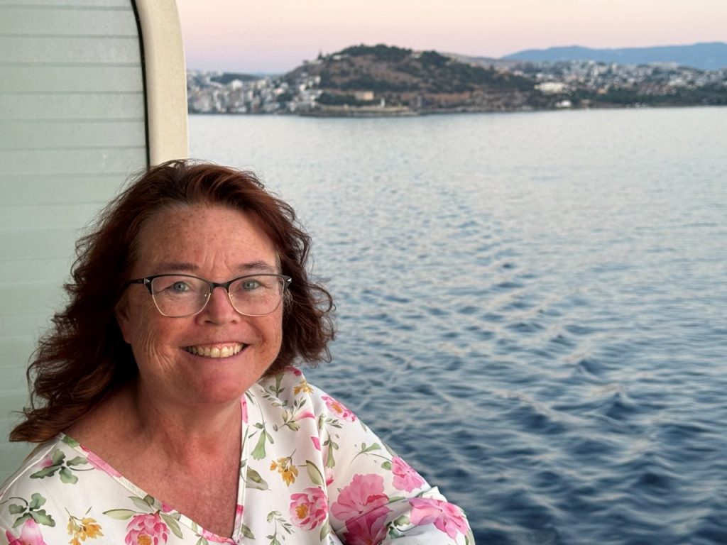 Kari smiles from the balcony of Celebrity Infinity wearing a white floral top, with calm blue water and the Greek coastline stretching behind her in the soft evening light. Her lightweight top was comfortable for relaxing outside on the ship after a hot day of exploring.
