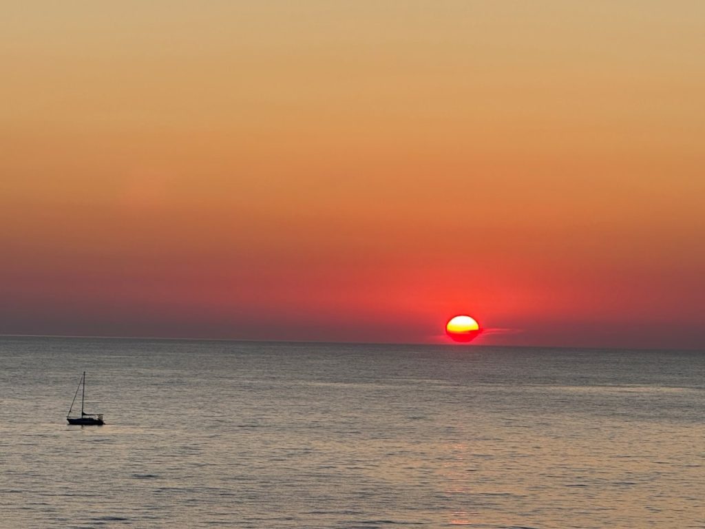 A glowing red-orange sun touches the horizon above calm water as the sky fades from gold to dusky pink. The peaceful view at sea captures the easy cruise experience that made sampling several parts of Greece work so well for Kari and Bill’s first visit.