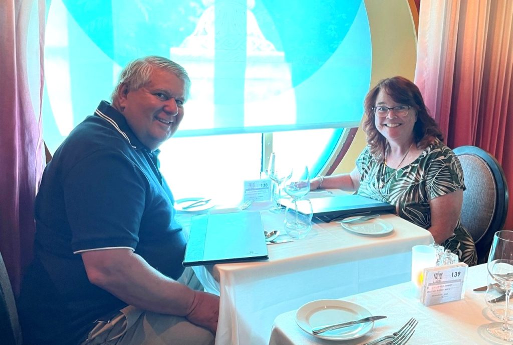 Bill and Kari sit together at a white-tablecloth restaurant table aboard Celebrity Infinity, with wine glasses and the sea visible through the large window behind them. Their experience cruising with and without a cruise drink package helped them decide when paying upfront is actually worthwhile.