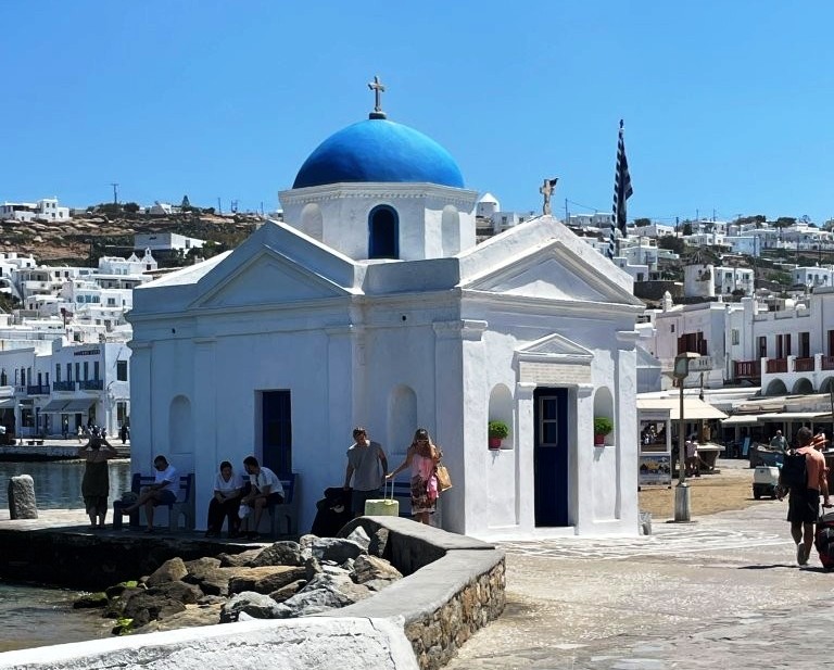 A small white Greek Orthodox church with a vivid blue dome stands beside the rocky Mykonos waterfront under a clear sky. Spending several nights on islands like Mykonos gives travelers more time for slower mornings, evenings and independent exploring.