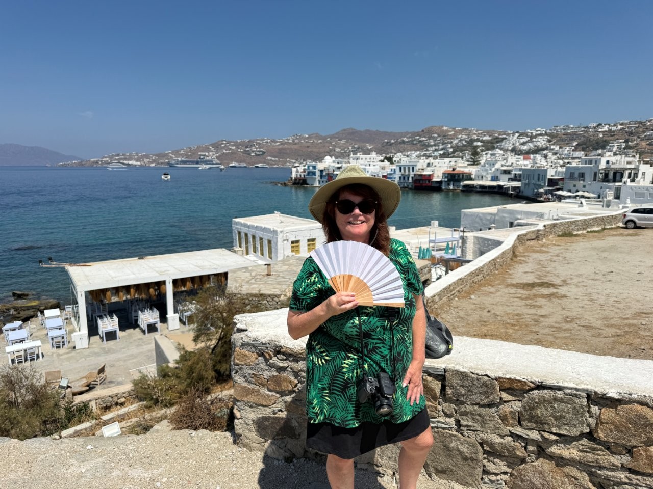 Kari stands above Mykonos harbor wearing a sunhat and holding an open hand fan, with white buildings, boats and blue water behind her. Exploring Mykonos independently during their cruise port day gave Kari a taste of the freedom she would have with several days on the island.