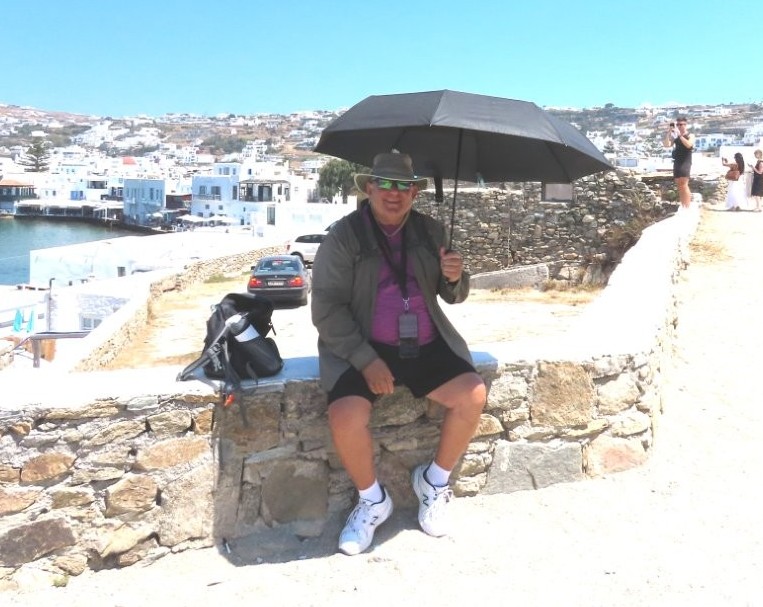 Bill sits on a stone wall in the hot sun with a large black umbrella opened to give him shade. His decision to bring an umbrella was a very smart "what to pack for a cruise" decision.