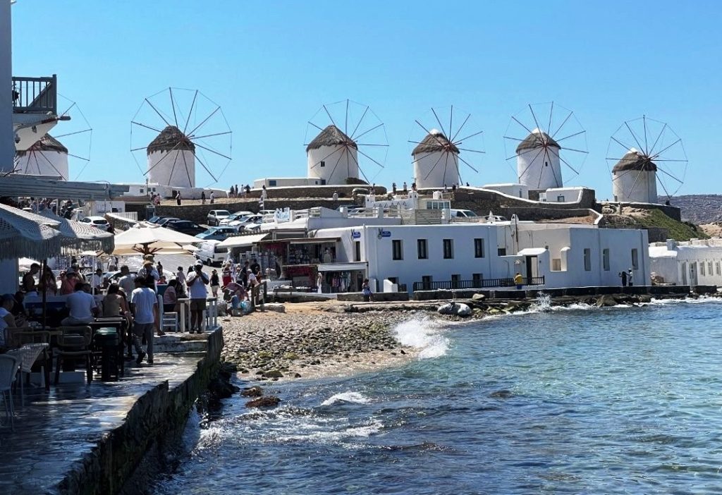 White cylindrical windmills topped with thatched roofs stand against a brilliant blue sky above Mykonos. The windmills, whitewashed lanes and blue water gave us the classic Greek-island scenery I had been waiting for.