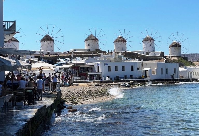White cylindrical windmills topped with thatched roofs stand against a brilliant blue sky above Mykonos. The windmills, whitewashed lanes and blue water gave us the classic Greek-island scenery I had been waiting for.