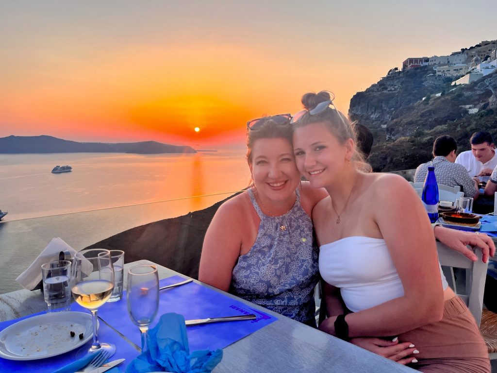 Melanie and her daughter smile together over dinner on a Santorini terrace as the orange sun sinks toward the Aegean Sea behind them. Staying overnight gave them time to enjoy Santorini at sunset and experience the island after many cruise passengers had returned to their ships.