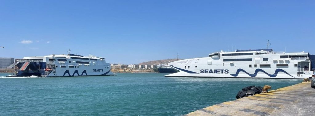 Large passenger ferries sit across the turquoise water at the busy port of Piraeus near Athens. The port represents the many connections and border-crossing logistics travelers may encounter while traveling through Greece and the Schengen Area.
