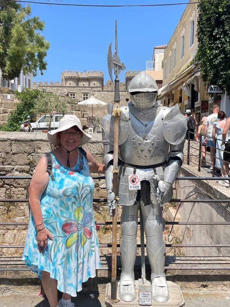 Kari stands beside a full suit of armor in Rhodes wearing a breezy summer dress, wide-brimmed sun hat and walking shoes. Her loose dress keeps her legs uncovered while her hat shades her face from the strong Greek sun.