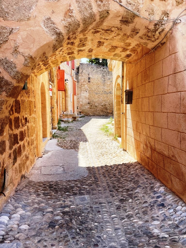A narrow stone passage leads through the medieval walls of Rhodes Old Town toward visitors walking in the bright sunlight beyond. Even while exploring historic areas like Rhodes, Kari and Bill found communicating in English surprisingly easy throughout their Greece trip.