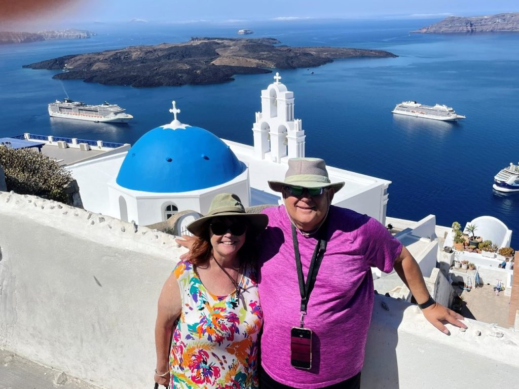 Kari and Bill stand beside the blue-domed Three Bells of Fira with the deep blue Aegean Sea stretching behind them in Santorini, Greece. Their lightweight summer clothes, hats and comfortable shoes are suited to a sunny day of sightseeing..