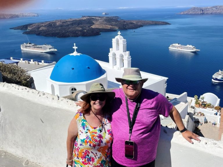Kari and Bill stand beside the blue-domed Three Bells of Fira with the deep blue Aegean Sea stretching behind them in Santorini, Greece. Their lightweight summer clothes, hats and comfortable shoes are suited to a sunny day of sightseeing..
