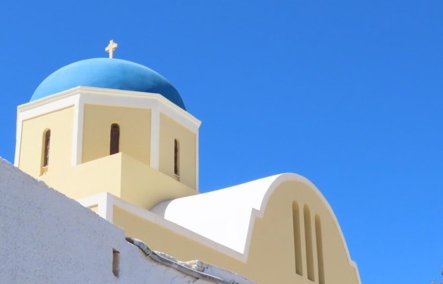 A bright blue church dome topped with a white cross rises above a cream-and-white building against a cloudless sky. Santorini’s blue-domed churches deliver the iconic Greek-island scenery many travelers hope to experience on a Celebrity Infinity Greece cruise.