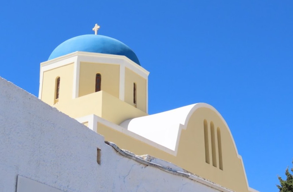 A bright blue church dome topped with a white cross rises above a cream-and-white building against a cloudless sky. Santorini’s blue-domed churches deliver the iconic Greek-island scenery many travelers hope to experience on a Celebrity Infinity Greece cruise.