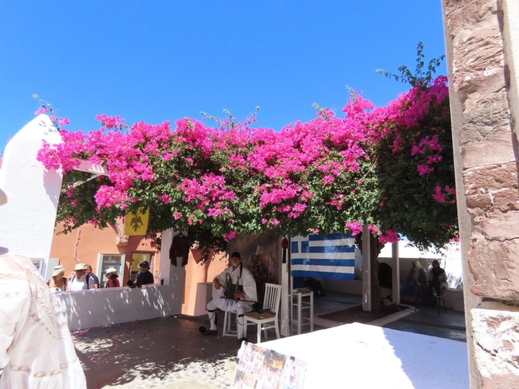Brilliant pink bougainvillea spills over a shaded whitewashed terrace in Santorini beneath a vivid blue sky. The colorful scene captures the Greek island atmosphere that draws so many travelers to Greece.