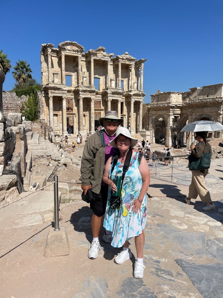 Kari and Bill stand in front of the towering stone façade of the Library of Celsus at the ancient ruins of Ephesus, Turkey. Both wear sun hats, lightweight clothes and comfortable walking shoes while exploring the sprawling archaeological site.