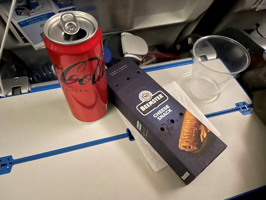 A red Coca-Cola can, a packaged chocolate snack, and a clear plastic cup sit on an open airplane tray table. It's amazing how something as simple as a cold drink and a snack can make a long journey feel a little more pleasant.