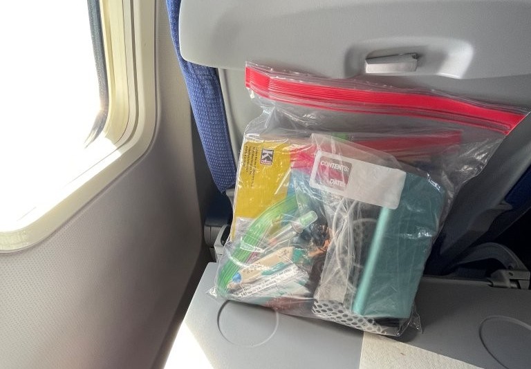 A transparent zip-top plastic bag filled with charging cords and small personal belongings rests beside an airplane window and seatback pocket. This is one of my favorite little travel habits because I feel much more settled when my personal essentials aren't scattered around me.