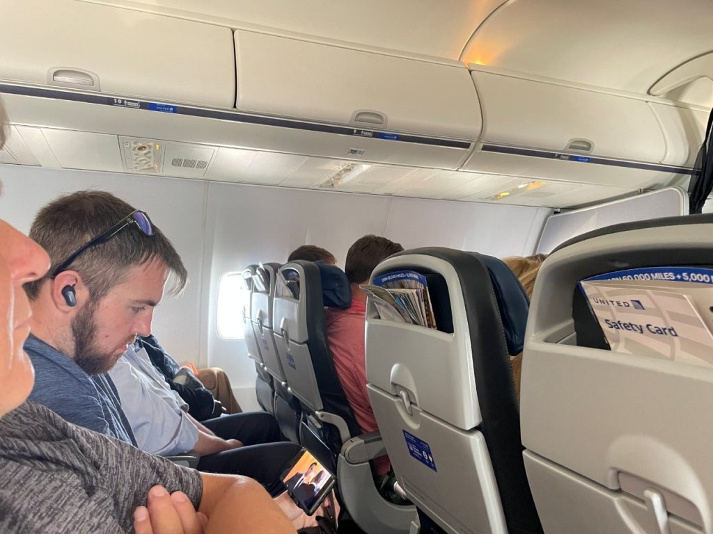 A man wearing an earbud looks at his phone while seated on an airplane, with several other passengers visible in the surrounding rows. Once the initial excitement of takeoff has passed, the cabin often settles into the familiar rhythm of people reading, resting, and entertaining themselves.