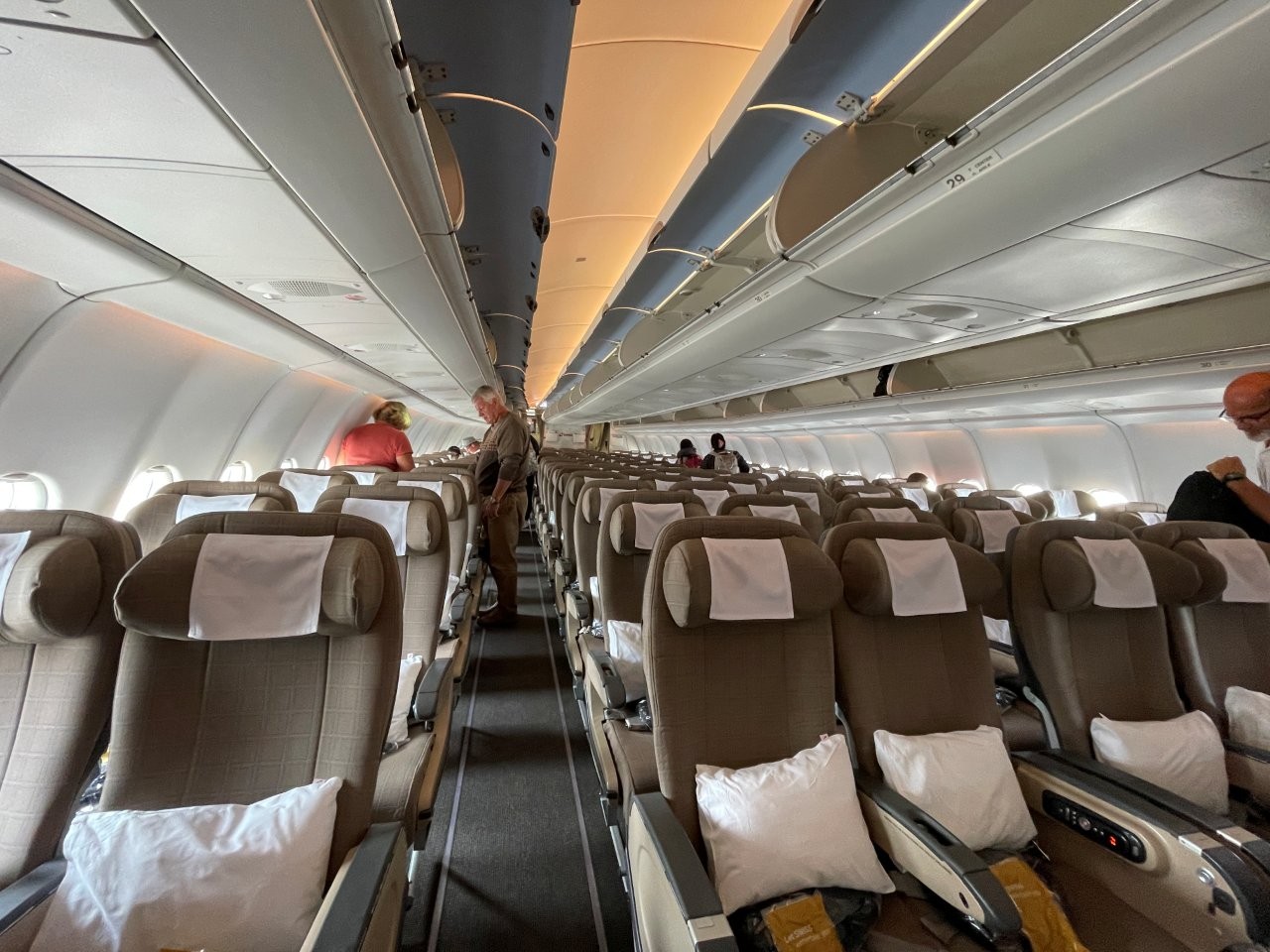 Wide beige airplane seats with white pillows and individual armrests line a central aisle beneath overhead luggage compartments. Bill and I generally choose Economy, but on exceptionally long flights, a little extra comfort can be worth the splurge.