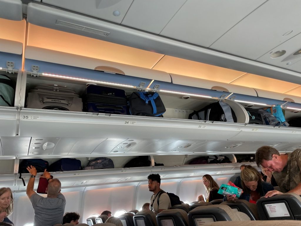 Open overhead bins are packed with carry-on suitcases and bags while passengers stand in the airplane aisle below. This chaos shows why it helps to stow your carry-on promptly and keep the smaller things you’ll need during the flight in your personal item.