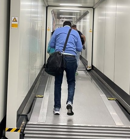 A traveler wearing a blue shirt and carrying a black shoulder bag walks down a long enclosed jetway with white walls, gray handrails, and overhead lights. The transition from a busy airport terminal to the airplane happens through a surprisingly ordinary-looking hallway.
