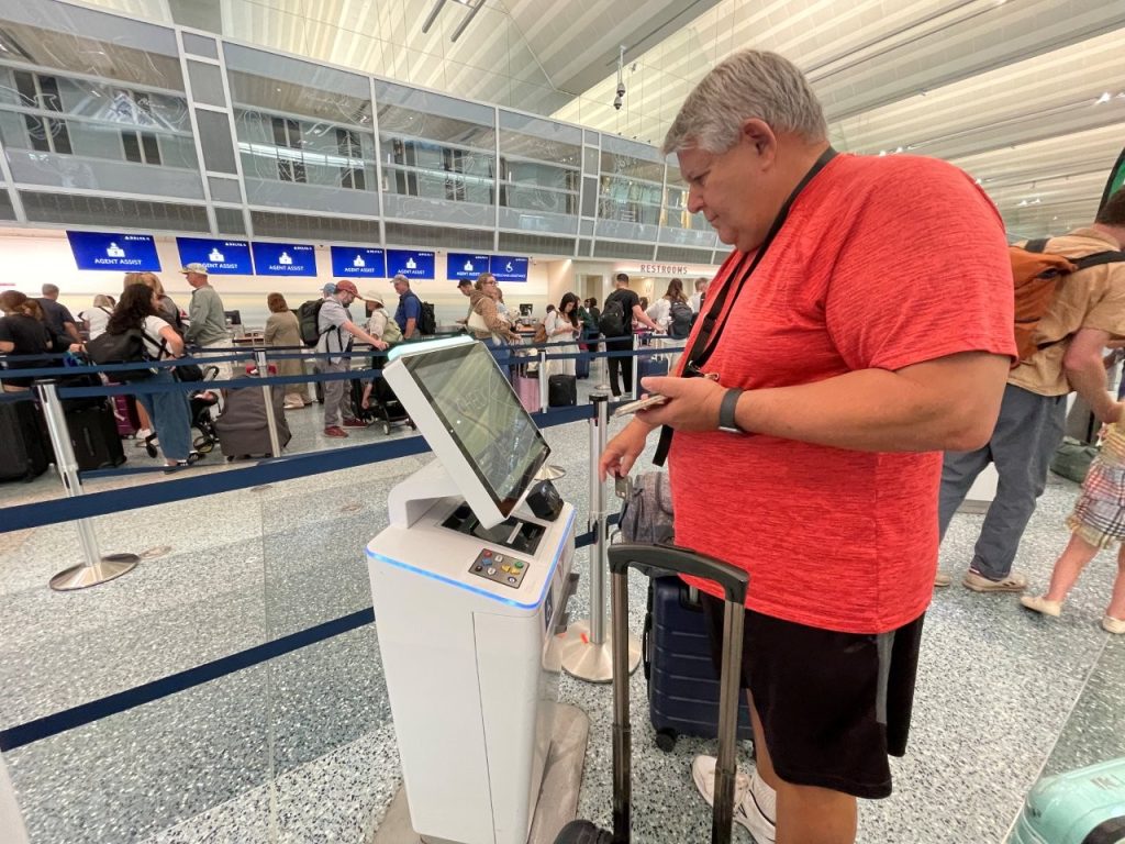 Bill, wearing a red shirt, operates a white touchscreen airport kiosk beside his suitcase while passengers wait at airline counters behind him. The first encounter with an unfamiliar airport machine can feel awkward, even when you're perfectly comfortable using technology elsewhere.