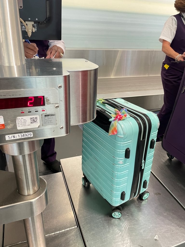 A turquoise hard-shell suitcase with a colorful luggage tag sits on a stainless-steel airport scale beside a red digital weight display. For someone flying for the first time, seeing the check-in process in action can make the airport feel less mysterious.