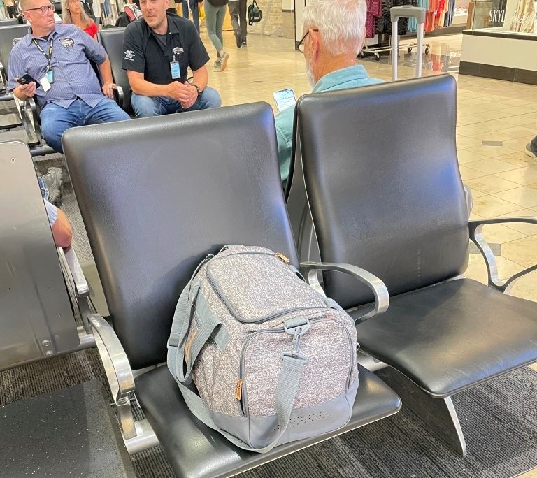 A gray duffle bag sits alone on a black airport waiting-area chair while several travelers talk in nearby seats. With so much activity happening around you, it's surprisingly easy to become distracted and lose track of your belongings.