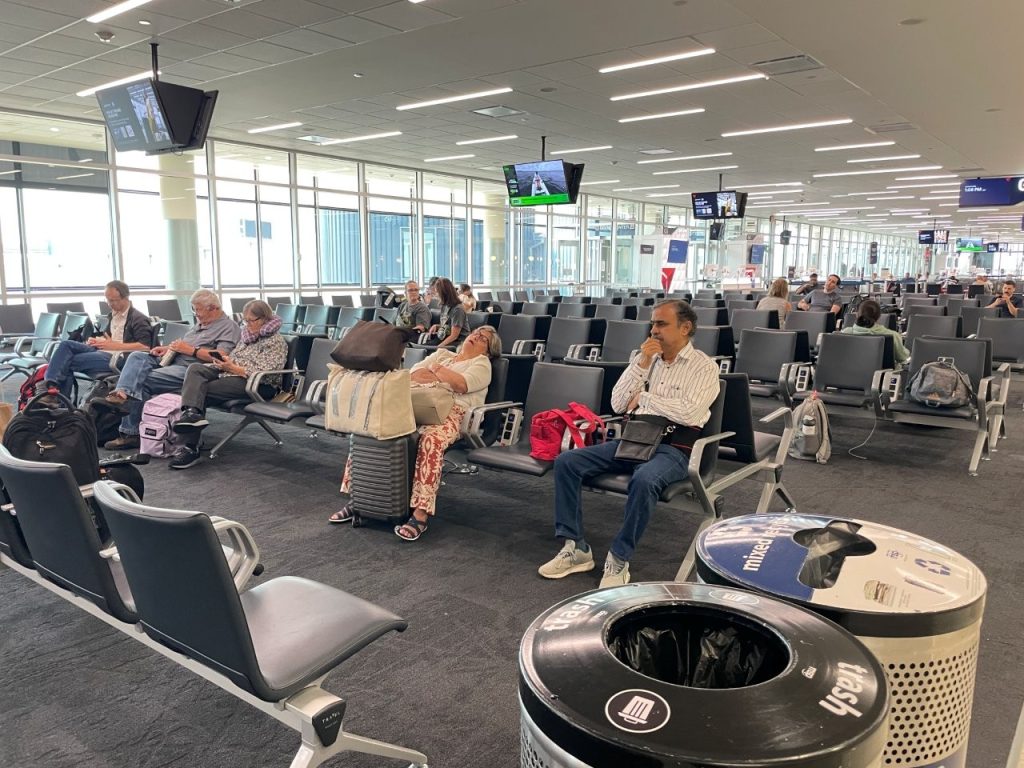 Travelers sit among rows of black airport chairs with suitcases and backpacks nearby, beneath overhead monitors and bright terminal lighting. After the activity of checking in and passing through security, this is often the first chance to settle down and enjoy a quieter moment before your flight.