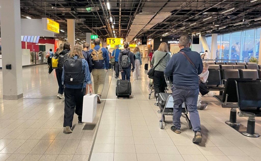 Travelers carrying backpacks and pulling wheeled suitcases walk through a busy airport corridor beneath an exposed ceiling and bright yellow directional signs. Large airports can feel like little cities, so it's reassuring to know that even experienced travelers sometimes need a moment to get their bearings.