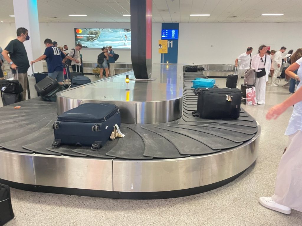 Black suitcases move along a wide stainless-steel airport baggage carousel while travelers stand nearby waiting for their belongings. Watching everyone reunite with their luggage is one of those ordinary airport moments that marks the end of a long journey.