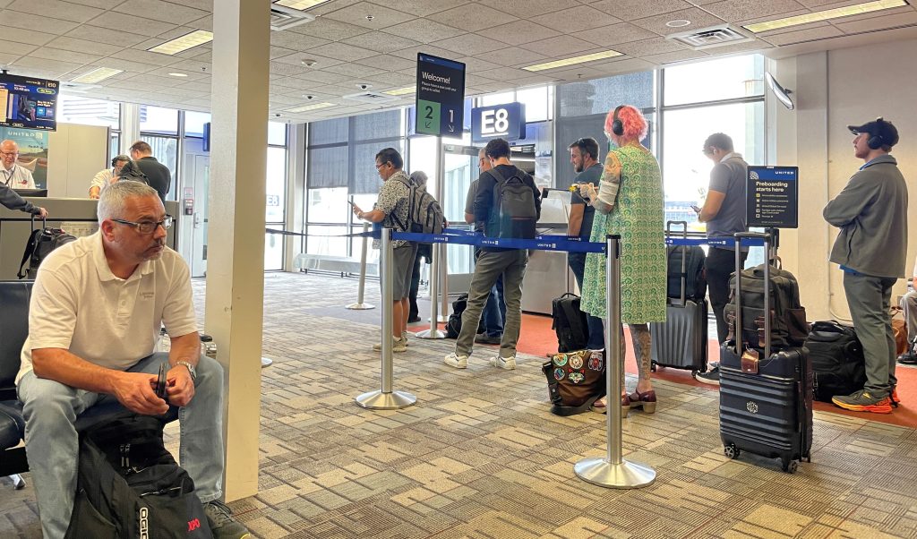 Travelers with rolling suitcases stand between queue barriers near airport gate E8 while other passengers wait in nearby seats. Watching an orderly boarding process can help take some of the uncertainty out of your first time flying.
