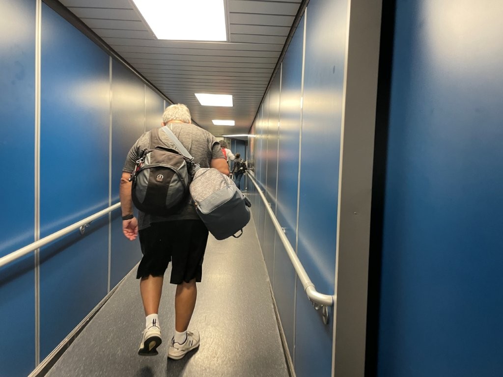 Bill walks down an enclosed airport jetway carrying a backpack and travel gear as he heads toward the plane. For someone flying for the first time, this is the moment after your boarding pass is scanned and you’re finally on your way onto the aircraft.