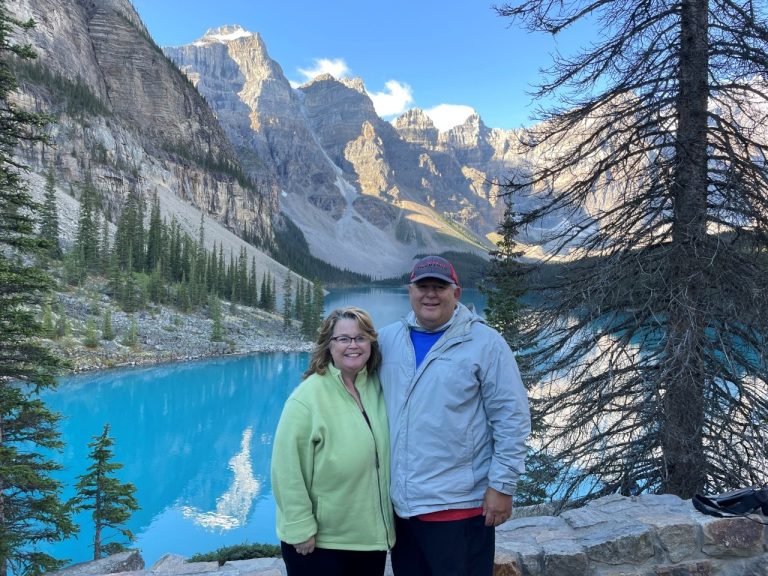 Kari & Bill stand in front of Moraine Lake at Banff National Park with snow-capped mountains in the background. Banff is one of our favorite places to travel.