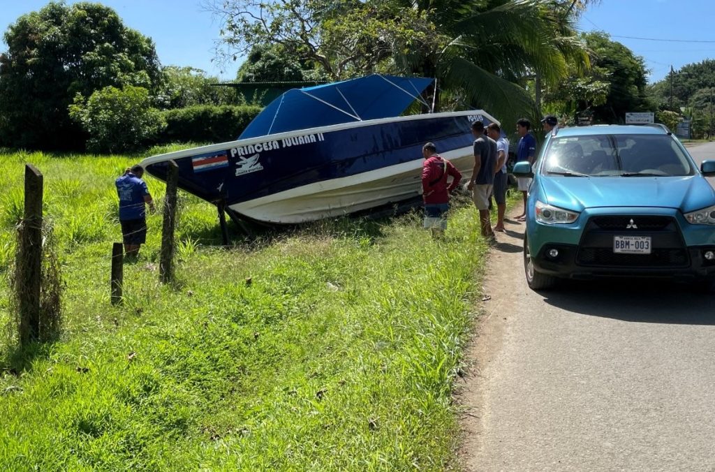 A long blue-and-white tour boat rests at an angle in tall grass beside a narrow road, with several people gathered nearby and concrete fence posts in the foreground. The photograph helps readers understand the position of the runaway boat and the fence that prevented Kari from escaping.