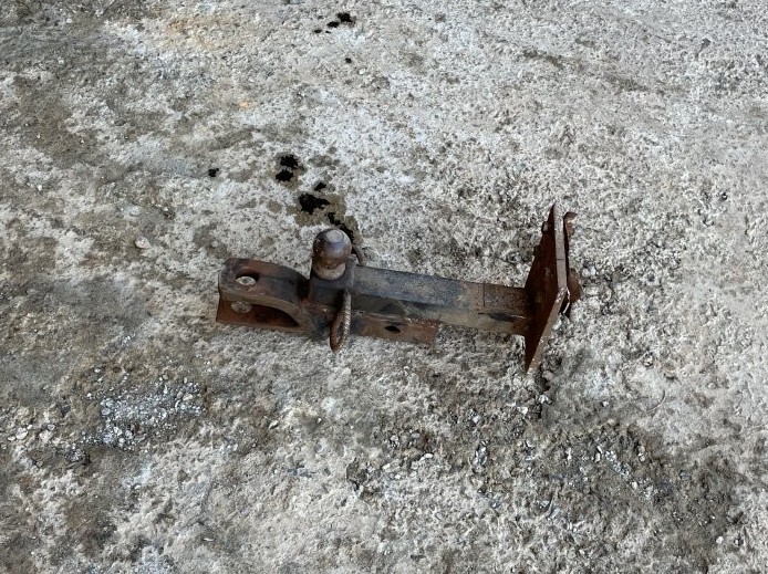 A detached, rusted metal trailer hitch lies on a rough concrete surface, with its tow ball and mounting hardware visible. This close-up shows the equipment involved in the frightening accident in Costa Rica.