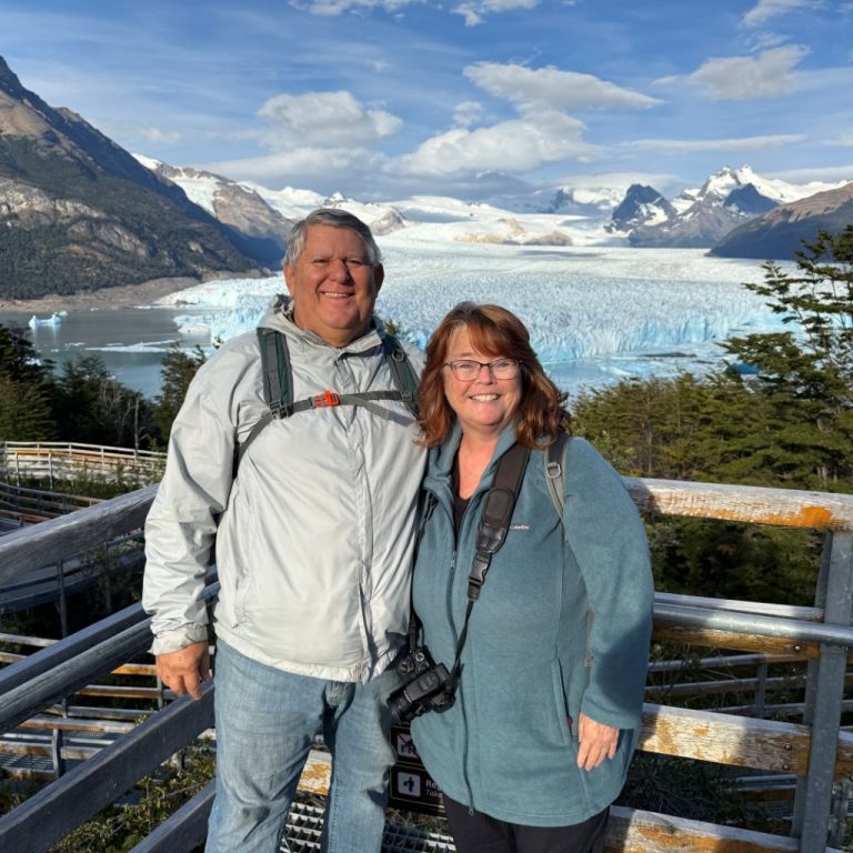 Kari and Bill stand together in front of a wooden fence with the massive Perito Moreno glacier behind them. Reaching Patagonia was one of the major experiences they checked off their travel bucket list.