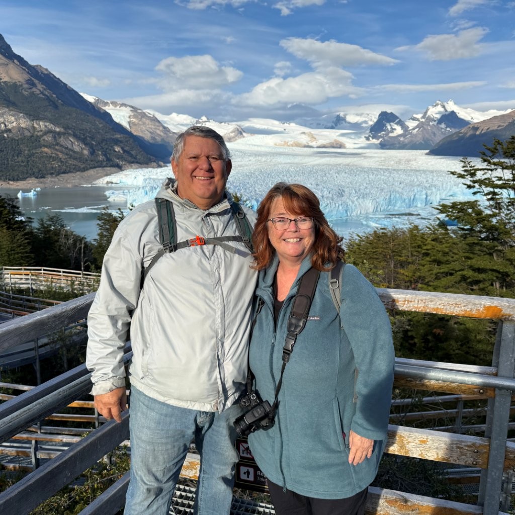 Kari and Bill stand together in front of a wooden fence with the massive Perito Moreno glacier behind them. Reaching Patagonia was one of the major experiences they checked off their travel bucket list.