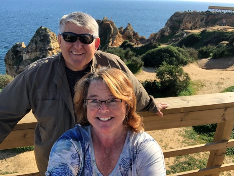 Kari and Bill smile for a selfie at a wooden overlook, with golden coastal cliffs, greenery, and the deep blue Atlantic Ocean behind them. This scenic stop highlights the beautiful coastal landscapes they discovered while exploring Portugal.