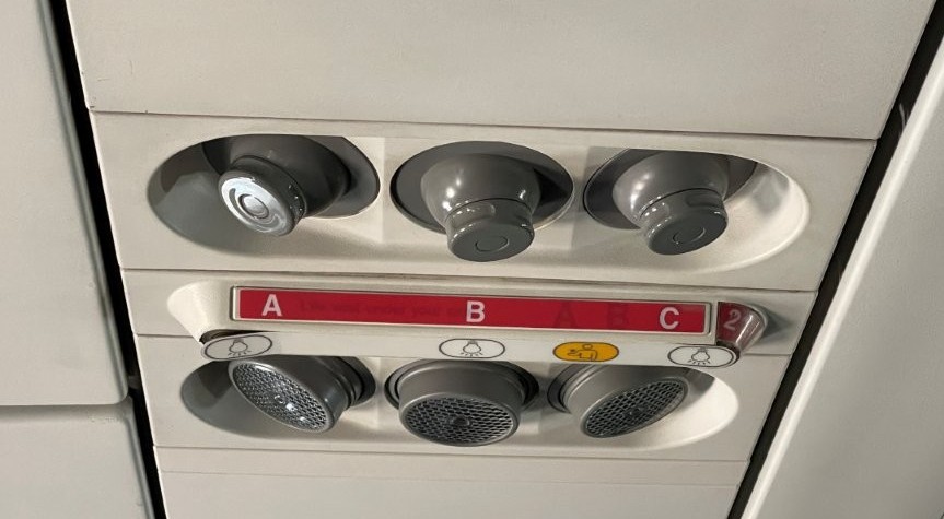 An overhead airplane panel displays red letters A, B, and C between circular air vents, reading lights, and small controls. Details like these are easy for frequent travelers to overlook, but recognizing them can give a first-time flyer a welcome sense of confidence.