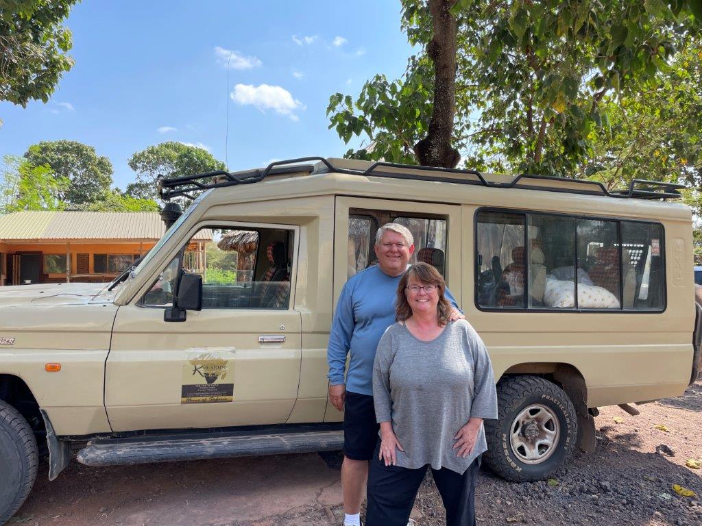 Kari and Bill stand beside a beige safari Land Cruiser parked beneath leafy trees, with the vehicle's pop-up roof visible above them. Their African safari fulfilled a major travel bucket list dream and strengthened Kari's passion for wildlife photography.