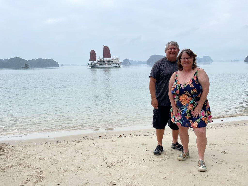 Kari and Bill stand on a sandy beach beside calm pale-blue water, with a traditional-style sightseeing boat and hazy limestone islands in the distance. Their visit to Ha Long Bay highlights the dramatic landscapes and coastal destinations they've experienced in Vietnam.