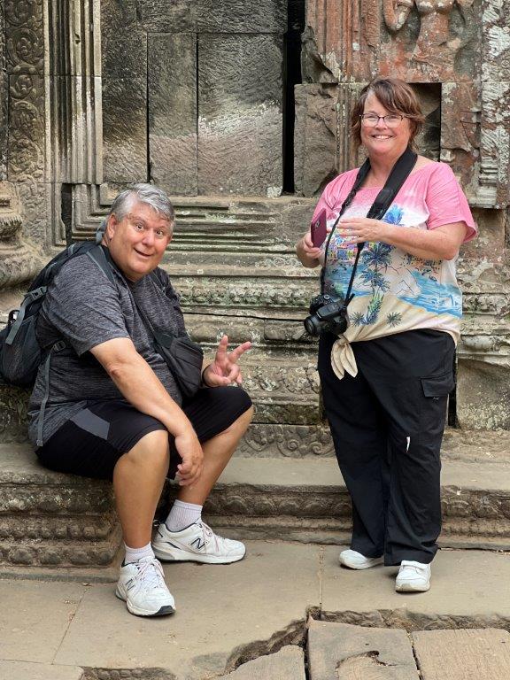 Bill sits on a stone ledge while Kari stands nearby, surrounded by weathered stone walls and intricately carved architectural details at Ta Prohm Temple. Exploring Cambodia's ancient temples added another fascinating cultural experience to their travels through Asia.